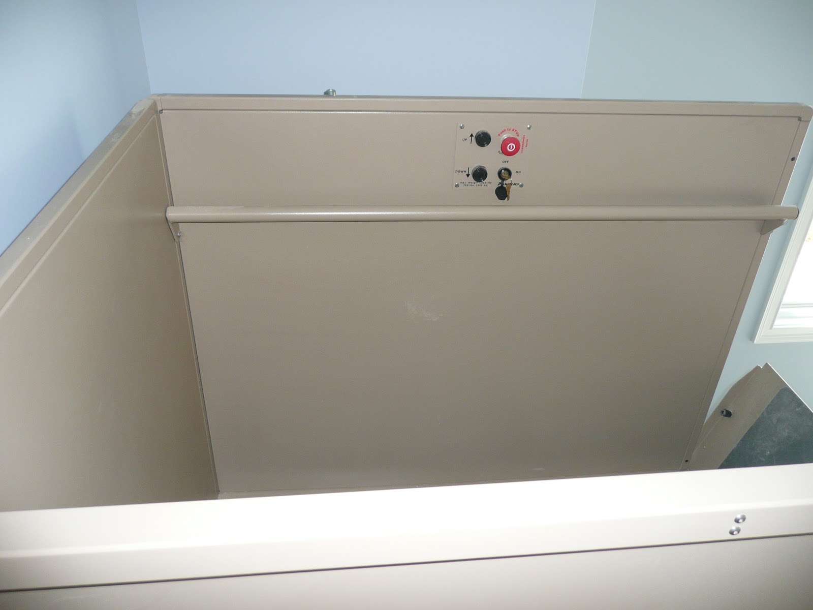 Interior view of a beige metal platform lift featuring a control panel with a red emergency stop button and key switch.