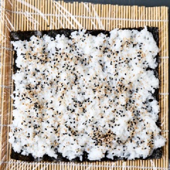 Bọc tấm tre cuộn cơm bằng màng bọc thực phẩm. Trải tấm rong biển lên trên, sau đó bạn múc cơm cho lên miếng rong biển. Dàn cơm cho thật đều khắp. Rắc lên cơm một lớp mè rang (trộn đều cả mè đen và mè trắng để tạo màu sắc đẹp cho cơm).