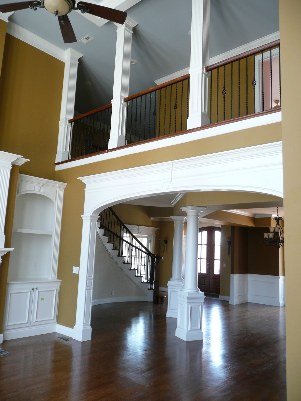 Interior view through a white arched opening showing hardwood floors, tan walls, white columns, and a second-story balcony with black metal railings.