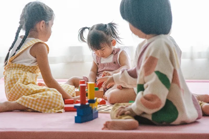 Three young children playing with colorful building blocks on pink mat in bright, minimalist room