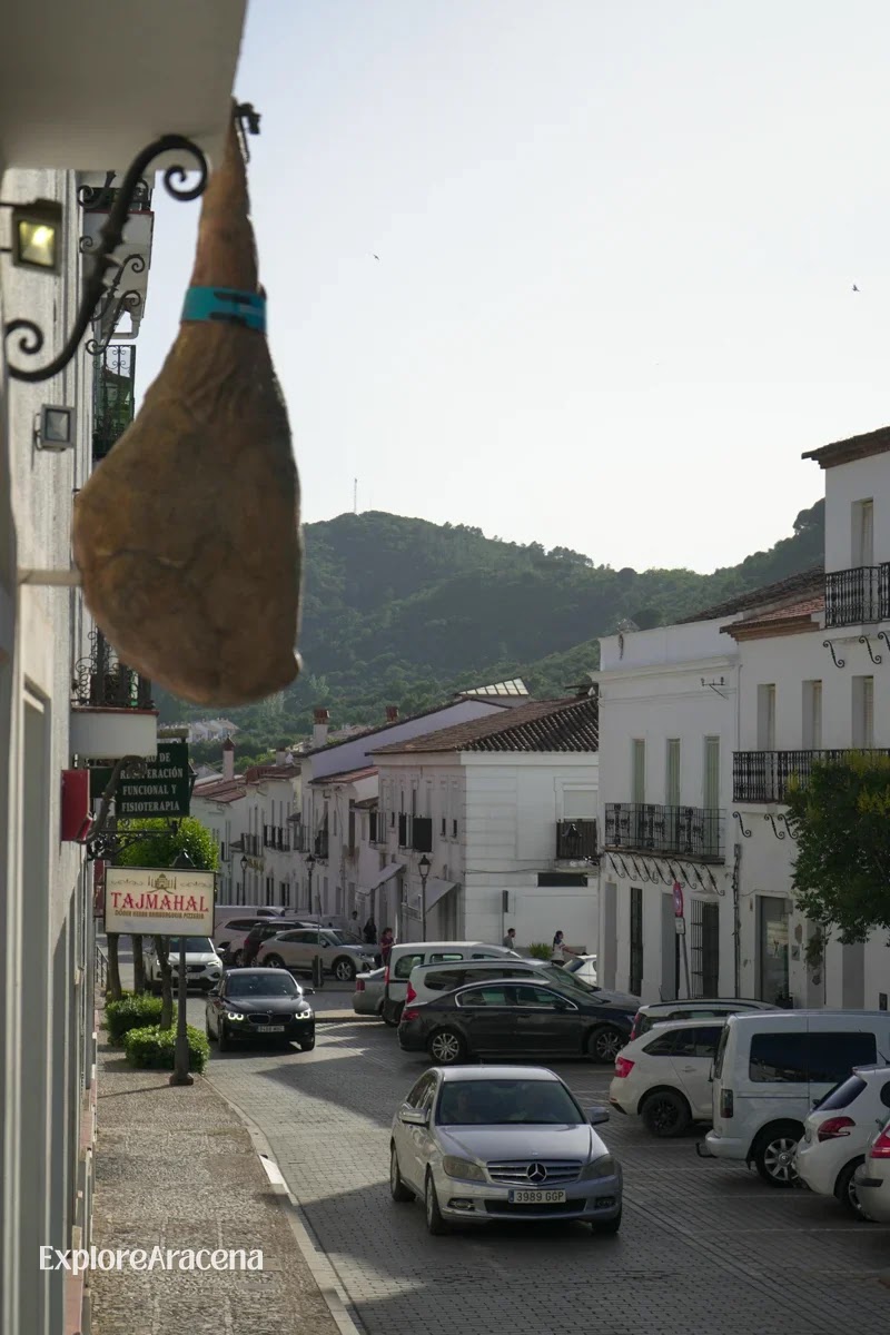 A leg of jamón hanging outside a shopfront on a street in Aracena, with white houses and the Sierra hills in the background