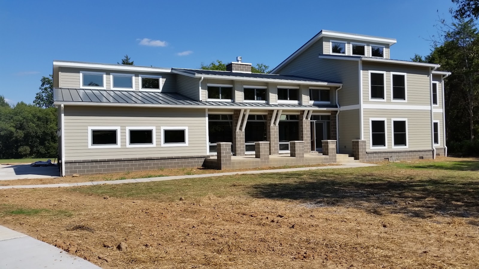 Exterior view of a multi-level custom home with tan siding, a metal roof, and numerous windows, featuring masonry block pillars and a dirt yard.