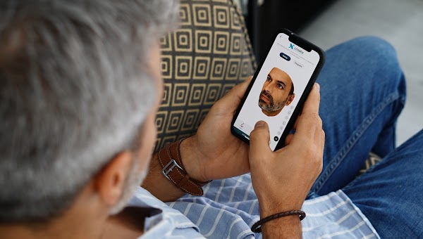 Photo prise de dos d’un patient masculin à domicile utilisant un smartphone pour visualiser une simulation 3D dans l’application Crisalix pour patients.