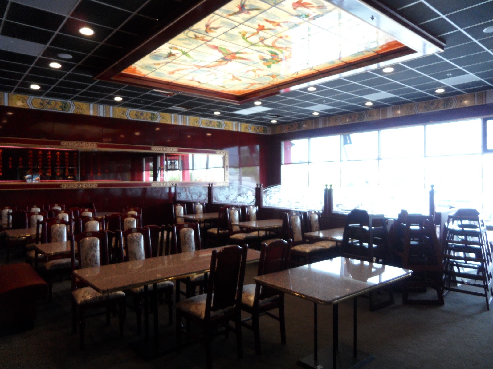 Restaurant dining room with rows of tables and chairs, featuring a large rectangular stained-glass ceiling panel and large windows along one wall.