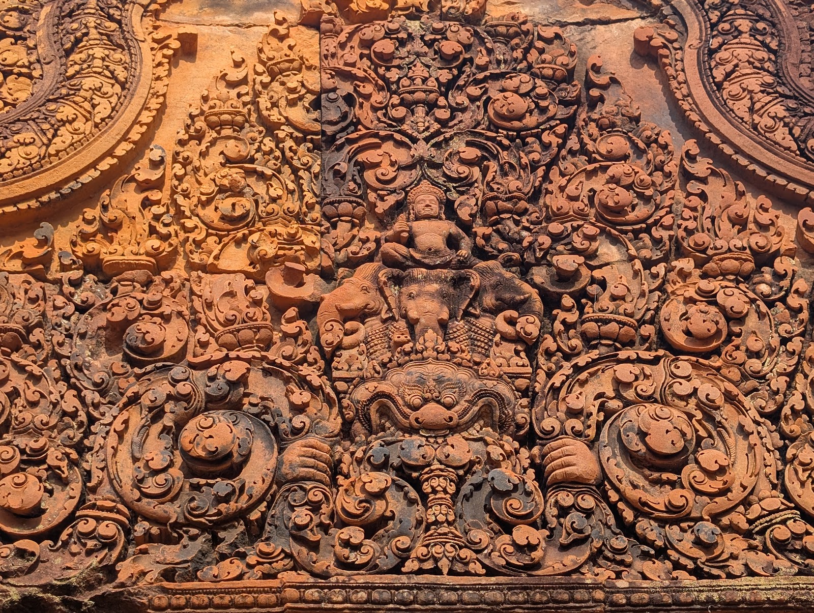 A complex red and orange bas relief showing deeply carved
                 floral patterns and a human figure on top of elephants on top
                 of a mask with hands gripping some of the flora to the sides