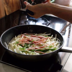 Phi thơm tỏi băm với 2 muỗng canh dầu ăn, cho khoai tây vào đảo đều 1-2 phút. Khi khoai tây hơi dịu xuống thì thêm vào ớt chuông và thịt xông khói. Nêm 1/4 muỗng cà phê muối, 1/4 muỗng cà phê tiêu, đảo đều cho tan gia vị. Khi khoai tây chín thì thêm 1 muỗng canh dầu mè vào, trộn đều rồi tắt bếp.