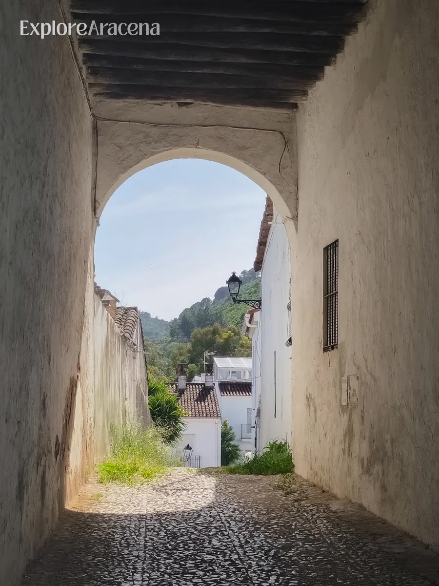 Alleyway in Aracena framed by an archway with white houses and green trees in the distance