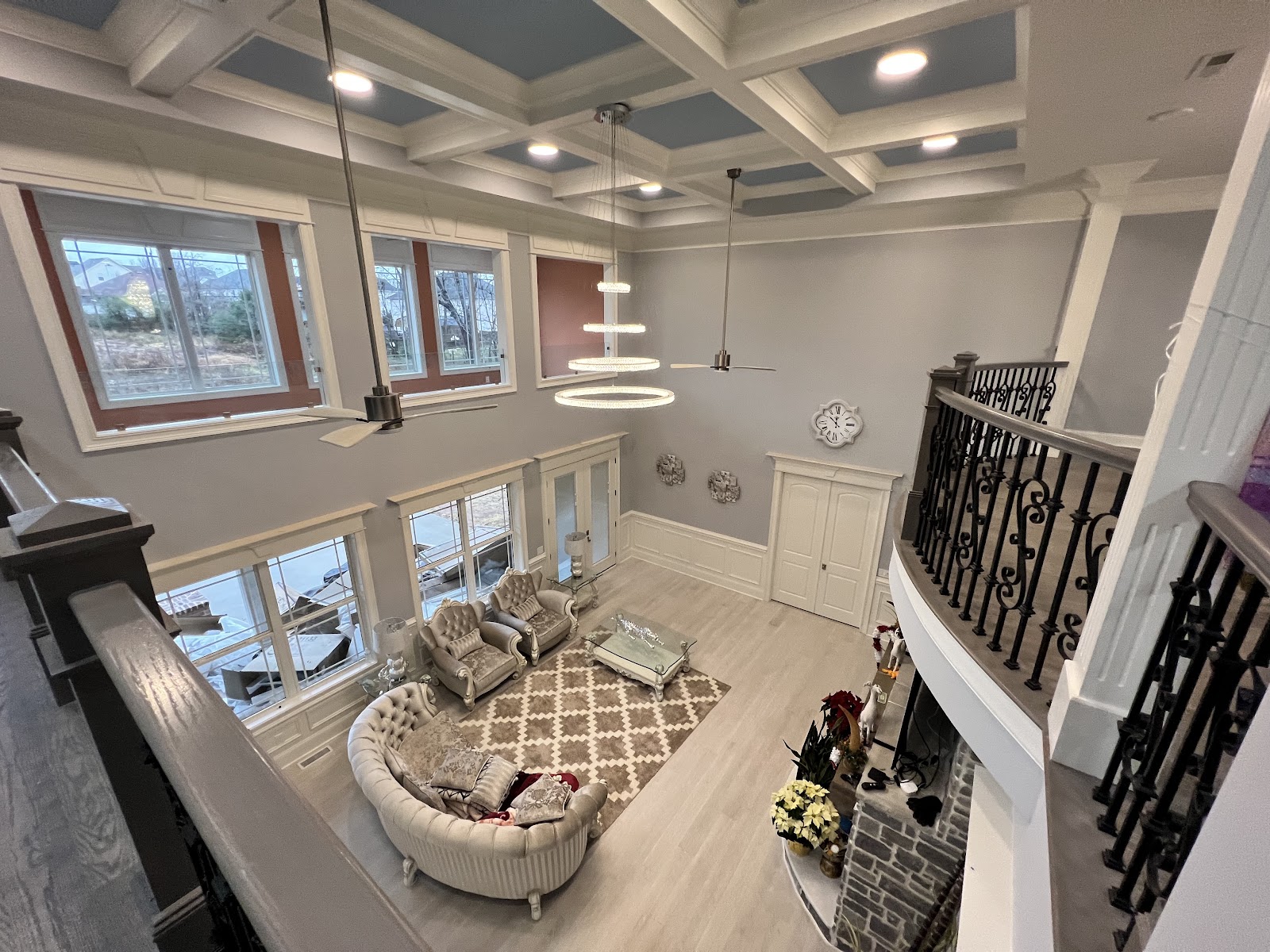 High-angle view of a living area featuring a coffered ceiling with blue panels, a tiered circular chandelier, a curved sofa on a patterned rug, and large windows.
