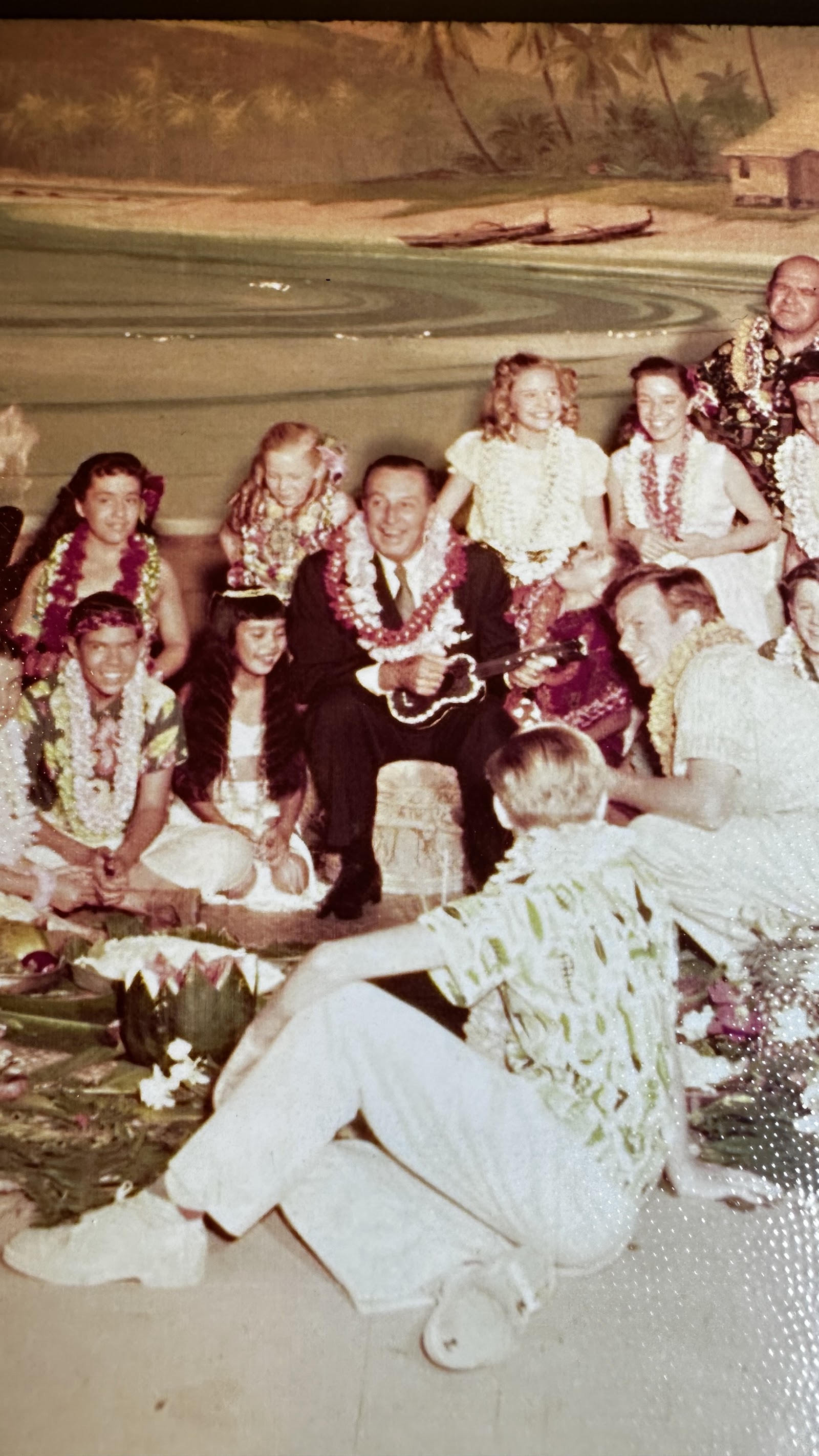 Vintage photograph of Walt Disney in a dark suit and floral lei, playing a ukulele while seated among adults and children in Hawaiian attire in front of a painted tropical backdrop, circa early 1960s