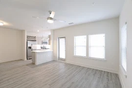 Open concept living room with wood finished flooring, ceiling fan, large windows, exterior door, and access to kitchen area