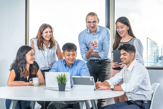 5 people in business attire gather around a table with a laptop and tablet