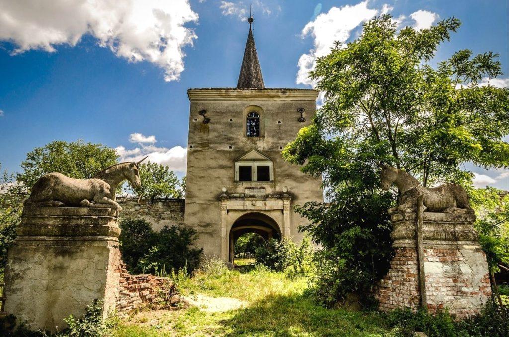 Kornis Castle, overgrown with plants and abandoned in Transylvania, Romania with two ancient unicorn statues in the foreground.