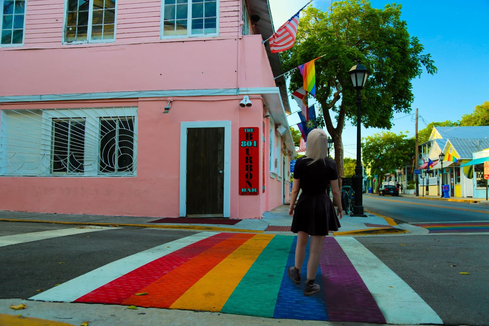 A person in all black is crossing a rainbow crosswalk. There is a pink building with a rainbow flag and across the street there is a bright yellow building with the sun shining on it (it also has a rainbow flag), and the rest of the image is in shadow.