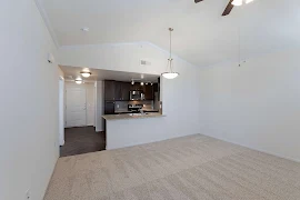 Living room with light walls and trim, neutral carpet & vaulted ceiling with a ceiling fan with a view to the kitchen & entry