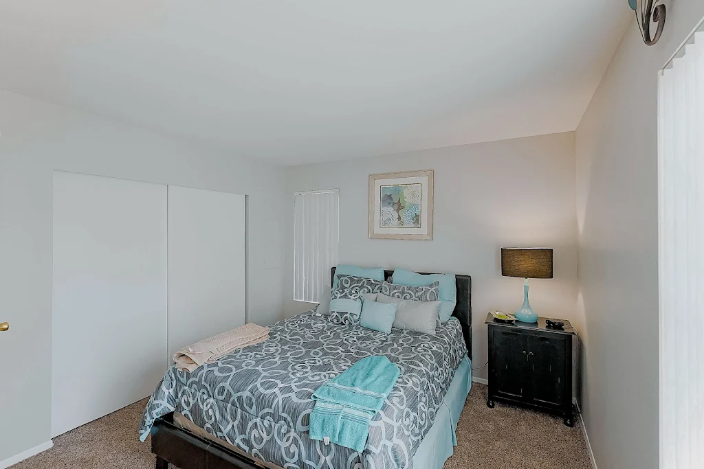 Model bedroom with gray patterned comforter and light blue accents, side table with lamp, white walls, and plush carpet