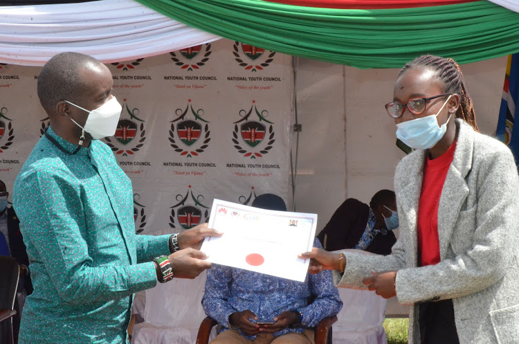 ICT Cabinet Secretary Joe Mucheru presents a certificate to one of the 100 youths who graduated after undergoing training in digital literacy at Karundas Primary School in Chaka, Nyeri, on Saturday.