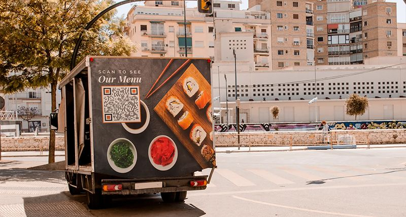 A food truck parked on the side of the road with a poster ad on back of the truck which has a conveniently placed QR code to access the menu. The poster also showcases sushi, sauces and chopsticks.