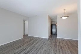 A bright, empty apartment interior with neutral walls, wood-style flooring, and a hanging light fixture in the dining area.