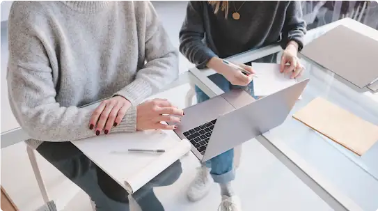 Duas pessoas trabalhando sentadas lado a lado em uma mesa de vidro com um laptop.