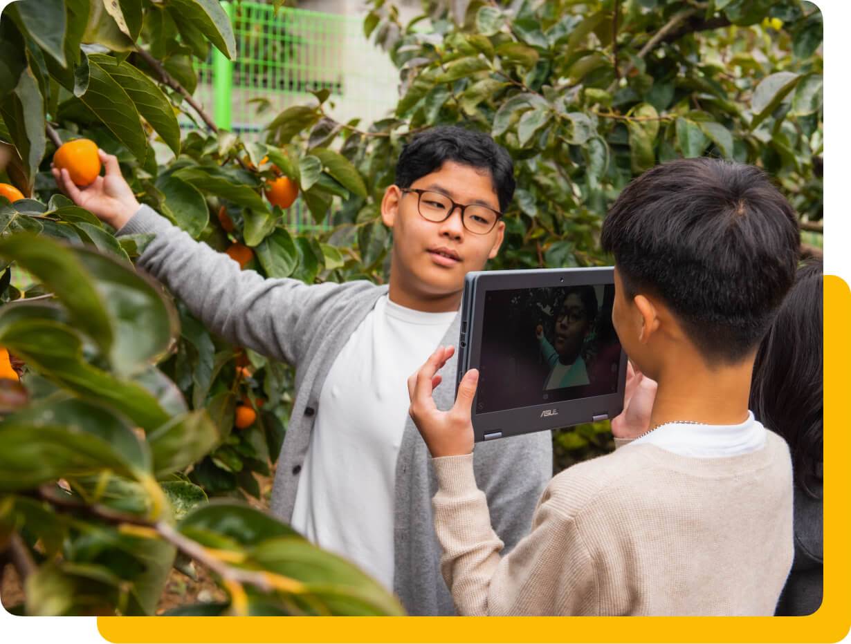 Dua orang pelajar lelaki sedang menggunakan Chromebook untuk mengambil gambar sebiji buah di taman.