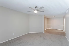 Empty living room in an apartment with beige walls, carpet flooring, a ceiling fan, and a dining area.