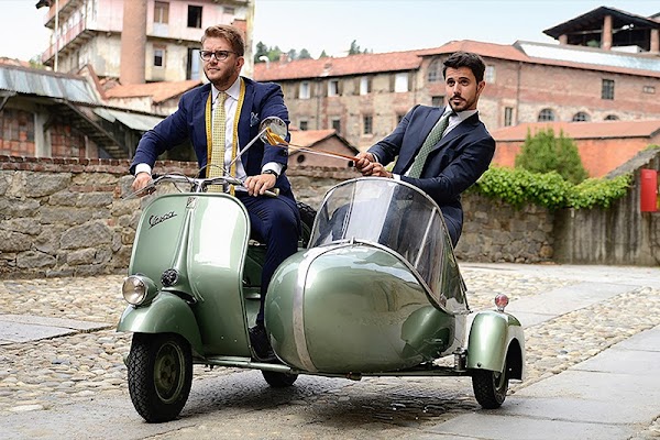 Due ragazzi vestiti elegantemente sfrecciano su una vespa con il sidecar all'interno di un borgo storico italiano