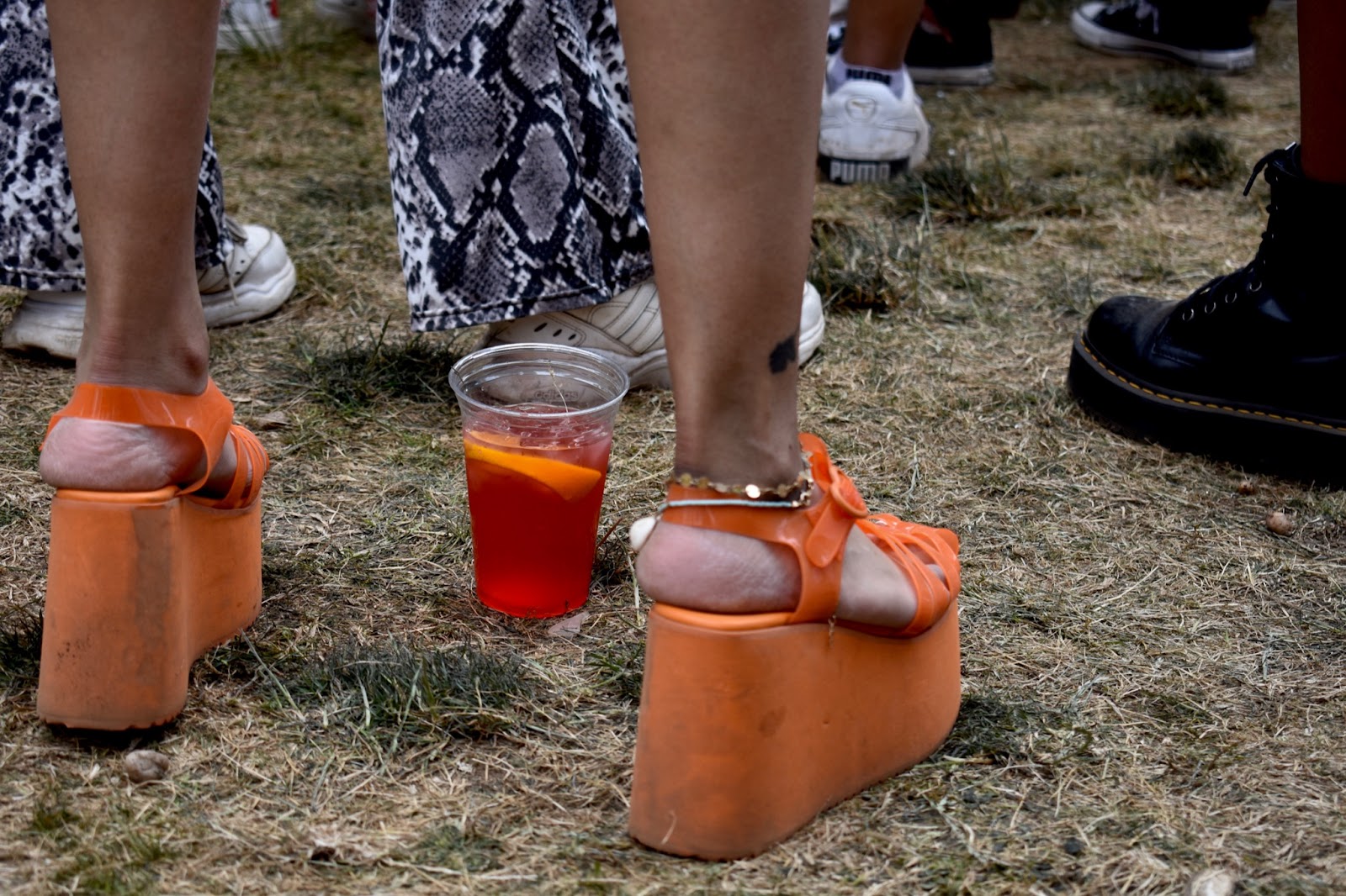 A woman’s feet in neon orange platform sandals complimented by a darker orange drink between her feet. 