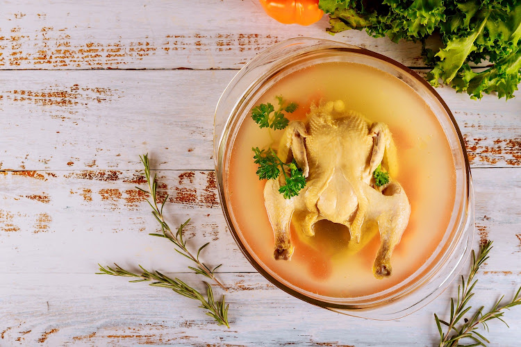 Chicken hot broth with carrots, whole chicken, onion, celery and parsley in glassl pot.