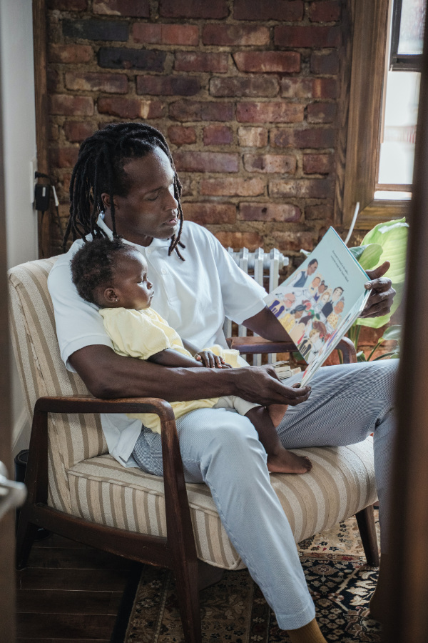 A young child sitting on their father's lap in an armchair as their father reads them a picture book.