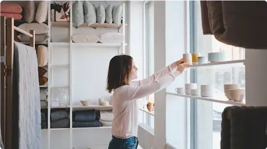 A business owner arranges products on a shelf in the front display window of her homegoods store.