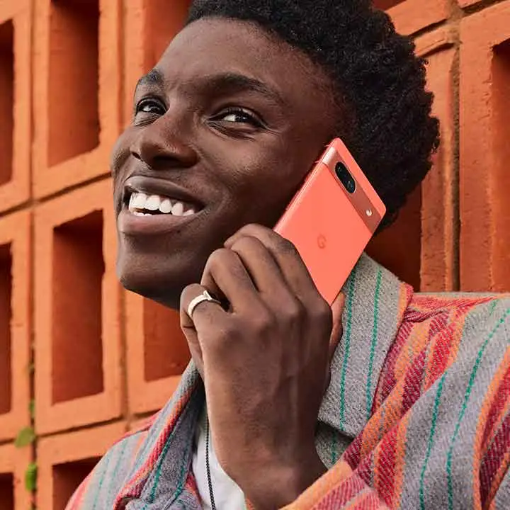 Person using Google Pixel 7a to take a call while smiling and leaning on an orange wall