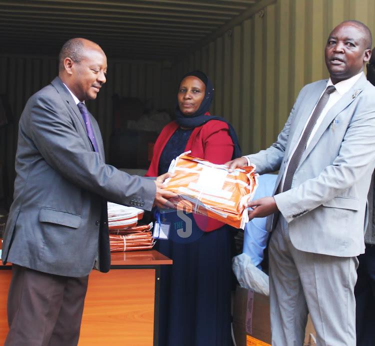 Kitui Education director John Thirigi hands over the KCSE exam papers to Kitui’s Muslim Secondary School principal Mohammed Radamshi at the deputy county commission’s office as Kitui Central subcounty Education director Mary Shano looks on on Friday, December 2