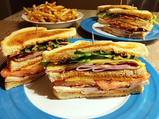 A club sandwich served on a blue plate with a bowl of French fries in the background