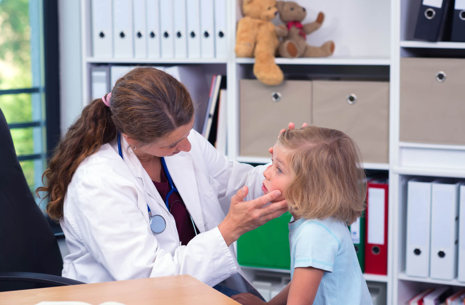 A female doctor that is sitting in her chair is closely examining the red eyes of a female child that is standing in front of her.