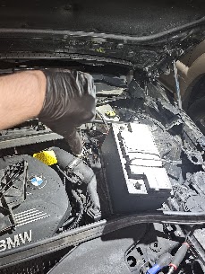 A mechanic wearing a black nitrile glove gives a thumbs-down gesture next to a car battery inside a BMW engine bay in Chicago.