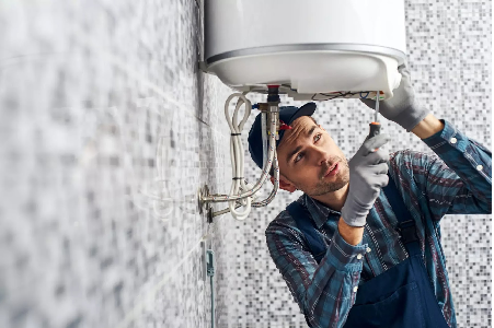 A plumber wearing a cap and overalls uses a screwdriver to repair a wall-mounted electric water heater tank in a bathroom.