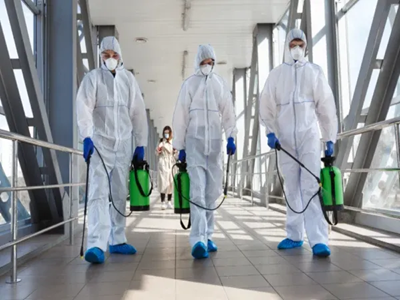 Three workers in white biohazard suits and respirator masks walking through an enclosed bridge walkway with green spray tanks.