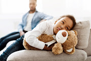 An upset little girl hugs a teddy bear, avoiding her father.