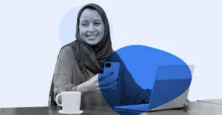 Una mujer con hijab sonríe mientras usa su smartphone y su portátil.