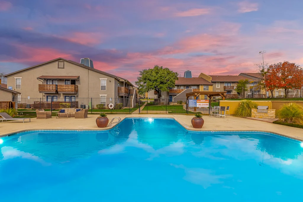 Grand Riviera's swimming pool at dusk with a BBQ grill, sundeck with seating areas and lounge chairs, next to buildings