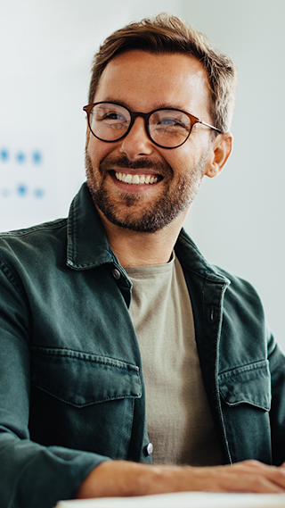 A white man with glasses is wearing a beige tee shirt under a green jacket smiling and working on his laptop while sitting in a conference room next to his Asian, female colleague wearing a white turtleneck and glasses.