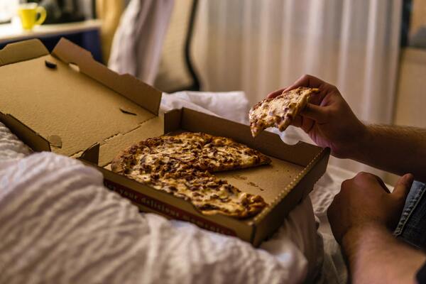 Foto de uma pessoa na cama com uma caixa de pizza no colo comendo