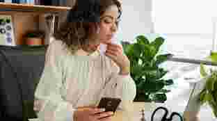 A woman seated at her desk, holding an Android-enabled phone and looking at a laptop.