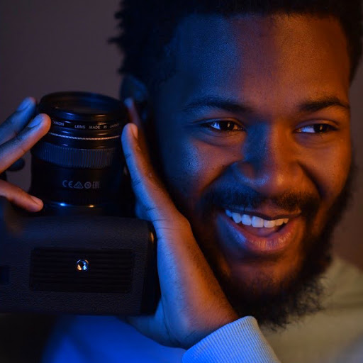 Portrait of artist holding their camera on their shoulder under colorful fluorescent lighting.