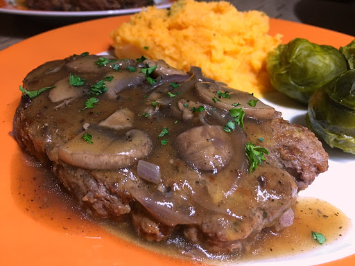 A steak topped with a mushroom sauce sitting on an orange plate with broccoli and mashed potatoes.