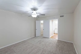 Bedroom with grey/tan walls, grey carpet, and a ceiling fan with grey blades with the doors to a walk in closet and hallway.