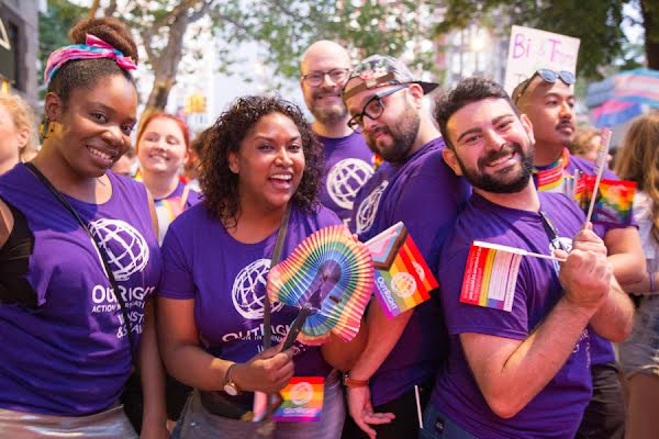Un grupo diverso de personas alegres en un evento al aire libre vistiendo camisetas moradas de Outright Action y ondeando banderas del arco iris del Orgullo Gay