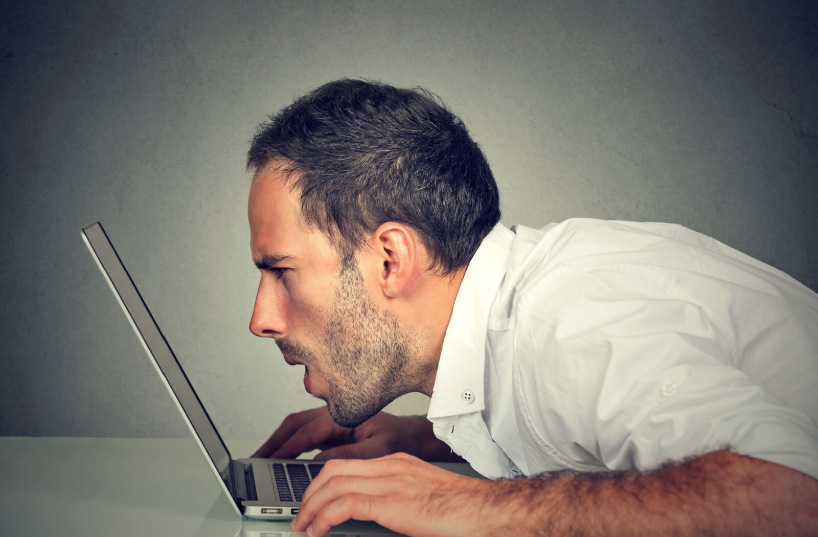 A man leaning forward and staring closely into a laptop with his mouth open wide and both hands resting on the desk.