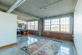 Apartment living room with cement floor, brick wall, large windows, and kitchen island in background