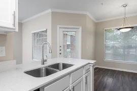 Modern apartment kitchen with white countertops, stainless steel sink, and wood flooring, leading to a living area with large windows.